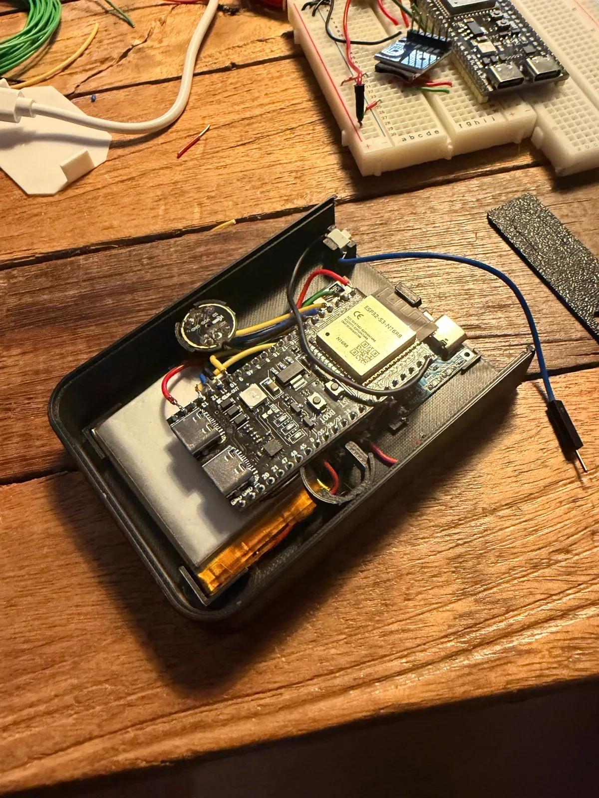 The open V1 shell on a wooden desk: an ESP32-S3 board, a round microphone and a LiPo battery wired inside a black 3D-printed case.