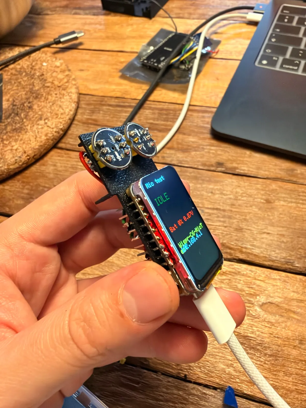 The V2 electronics held in a hand: two round microphones above a small board whose screen reads “Mic test, IDLE” with battery and network status.